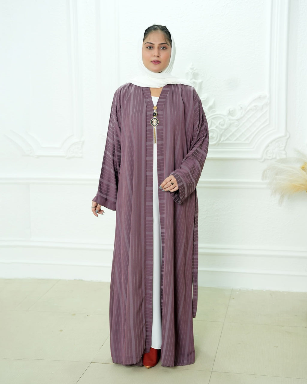 A woman wearing a long, loose-fitting mauve-purple abaya with vertical stripes over a white inner dress, a white hijab, and red shoes, standing in front of a white decorative wall.