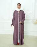 A woman wearing a long, loose-fitting mauve-purple abaya with vertical stripes over a white inner dress, a white hijab, and red shoes, standing in front of a white decorative wall.